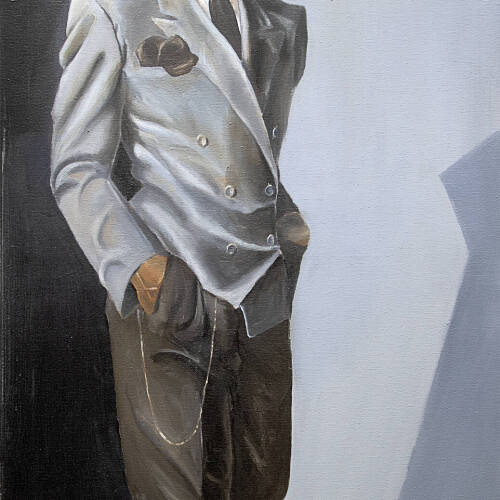 Man in a gray zoot suit wearing a hat with hands in his pockets. He is standing with one foot in front of the other.